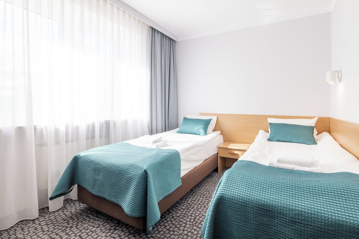 Una habitación de hotel con dos camas, una mesita de noche, una lámpara y una ventana con cortinas translúcidas.