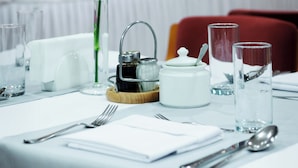 Una mesa de comedor con mantel blanco, un jarrón con una flor amarilla, una tetera, una taza, un vaso y cubiertos.