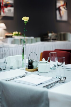 Una mesa de comedor con mantel blanco, un jarrón con una flor amarilla, una tetera, una taza, un vaso y cubiertos.