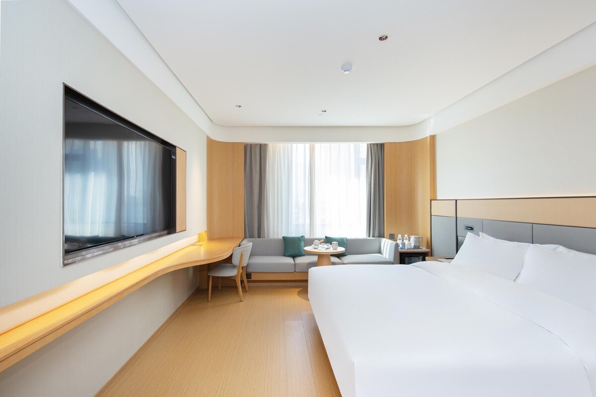 A modern hotel room with a large bed, a desk, a sofa, and a window with curtains.