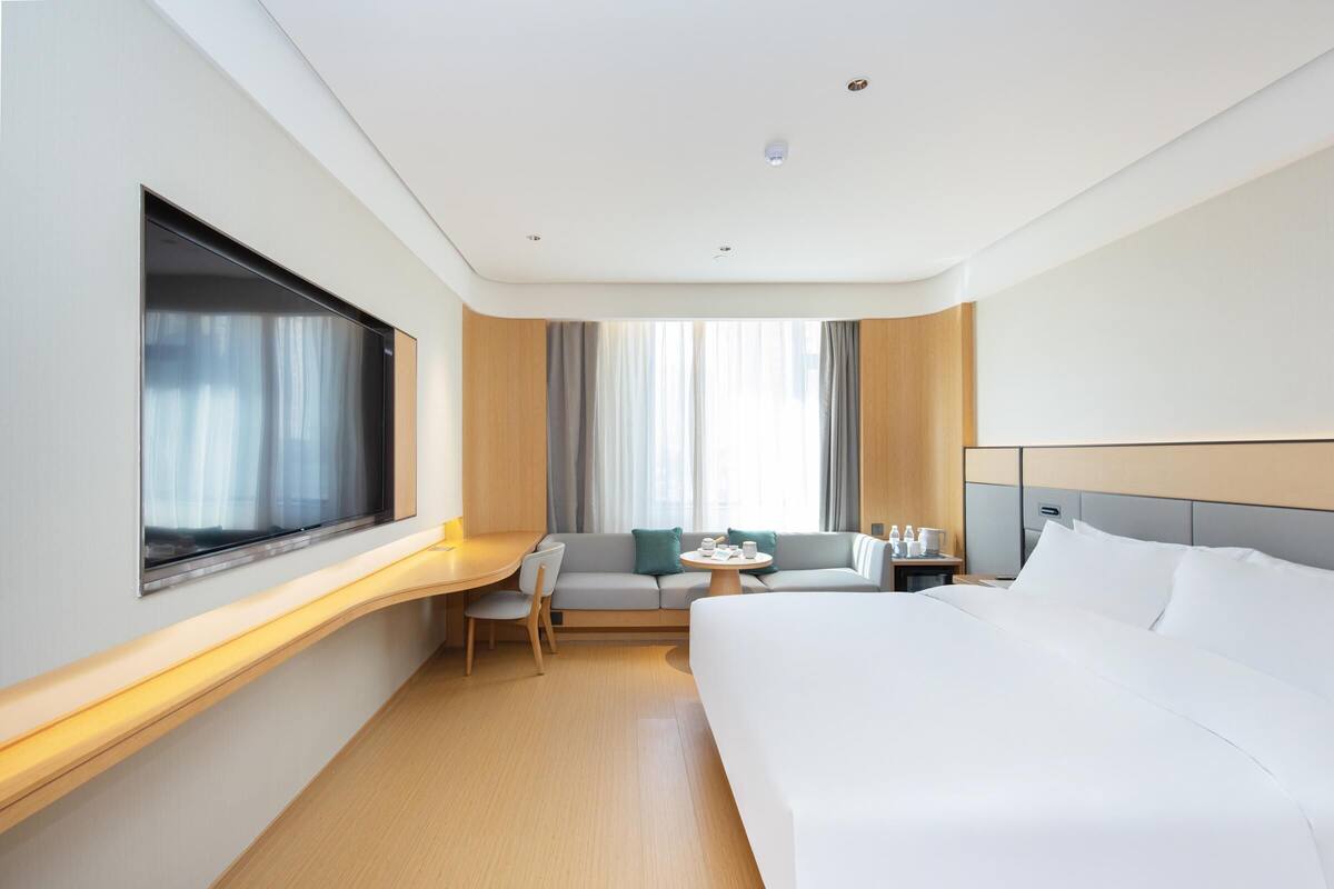 A modern hotel room with a large bed, a desk, a sofa, and a window with curtains.