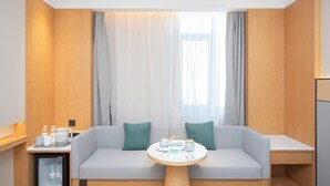 A modern hotel room with a sofa, a small table, a mini-fridge, and a window with curtains.