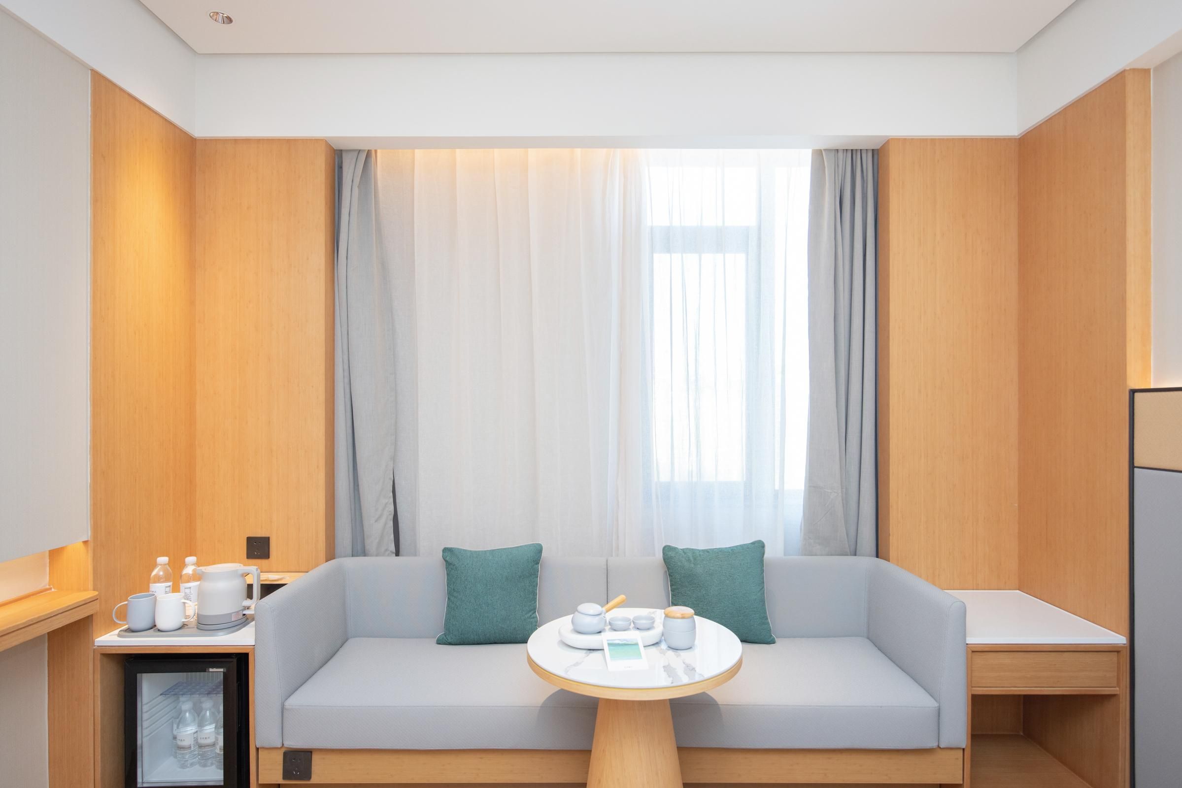 Una habitación de hotel moderna con sofá, una mesita, un mini refrigerador y una ventana con cortinas.