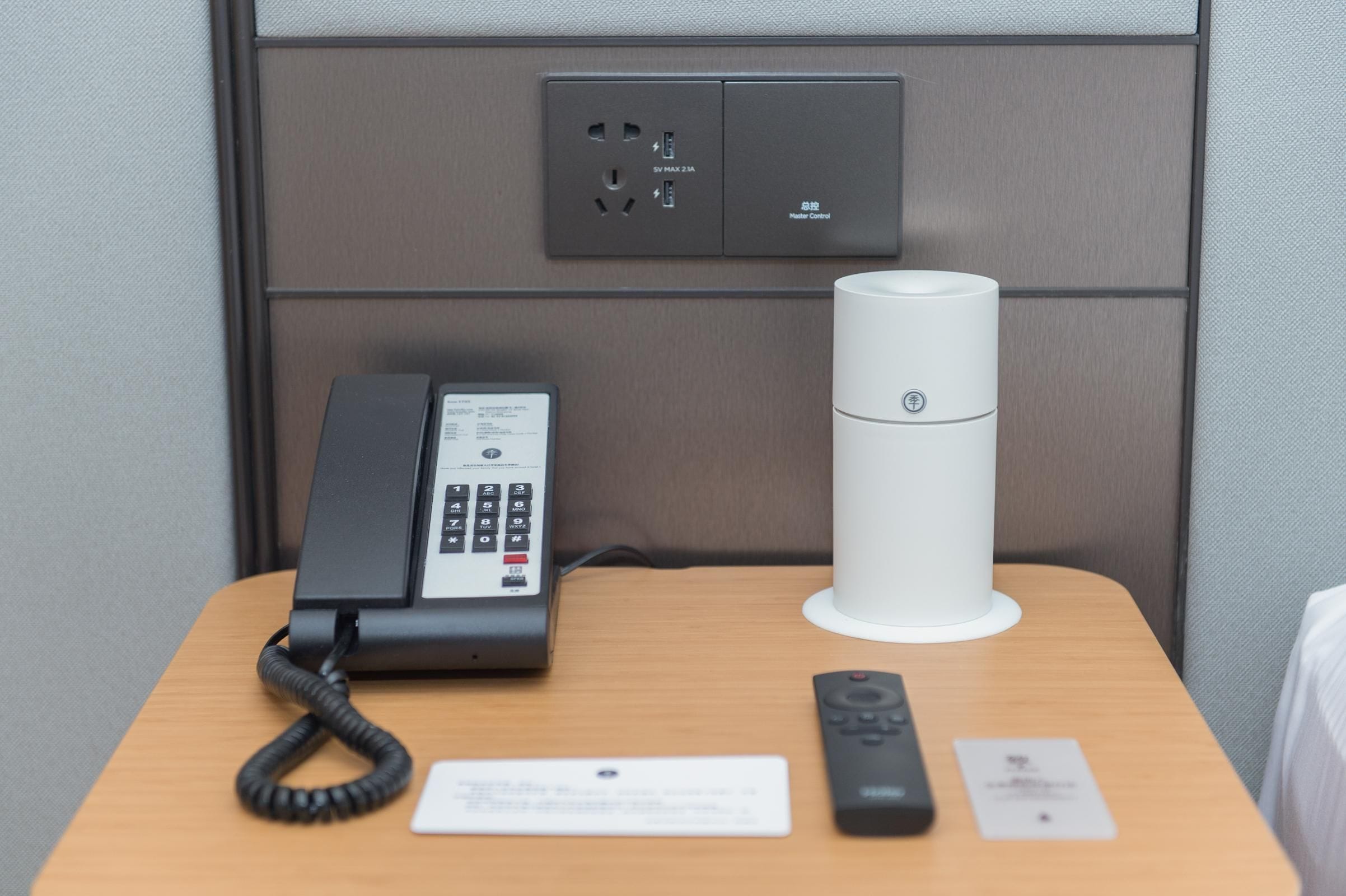 Quarto de hotel com telefone, um alto-falante branco e controle remoto sobre uma mesa de madeira.