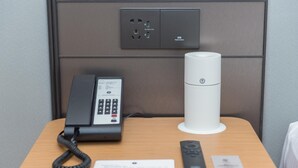 A hotel room with a desk, a telephone, a remote control, and a wall-mounted power outlet.