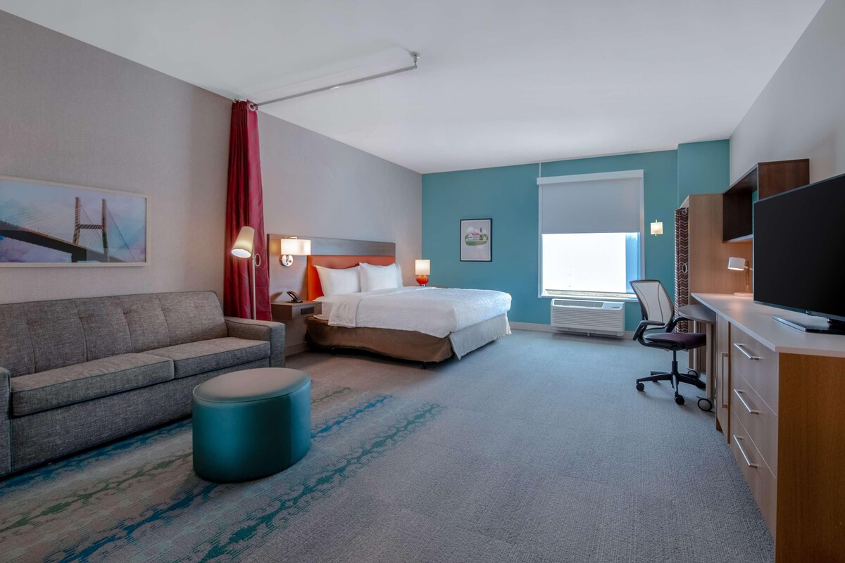 Une chambre d’hôtel avec un lit, un canapé, un bureau avec un ordinateur, une télévision et une fenêtre avec des rideaux.