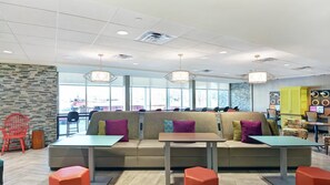 A modern cafe with a central seating area featuring a sofa and tables, large windows providing natural light, and a visible outdoor view. - Home2 Suites By Hilton Savannah I95 North (Port Wentworth)