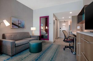 Quarto de hotel com sofá, um banquinho redondo, escrivaninha com cadeira, televisão e espelho com moldura rosa.