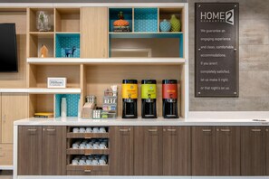 Une petite cuisine d’hôtel avec une pancarte indiquant « GARANTIE HOME 2 », une autre pancarte indiquant « HILTON HONORS » et trois machines à café étiquetées « GOURMET », « DÉCAFÉINÉ » et « EAU CHAUDE ».