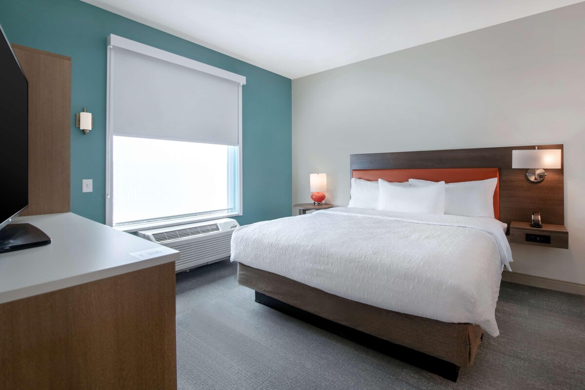 Une chambre d’hôtel avec un grand lit, un bureau, une télévision, une fenêtre avec des stores et une lampe fixée au mur.