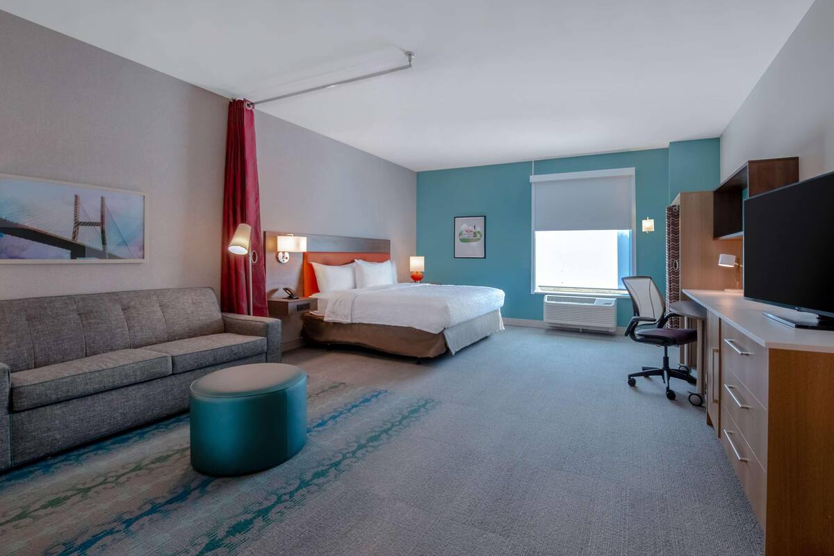 Habitació d'hotel amb llit, sofà, escriptori amb ordinador, televisió i finestra amb cortines.