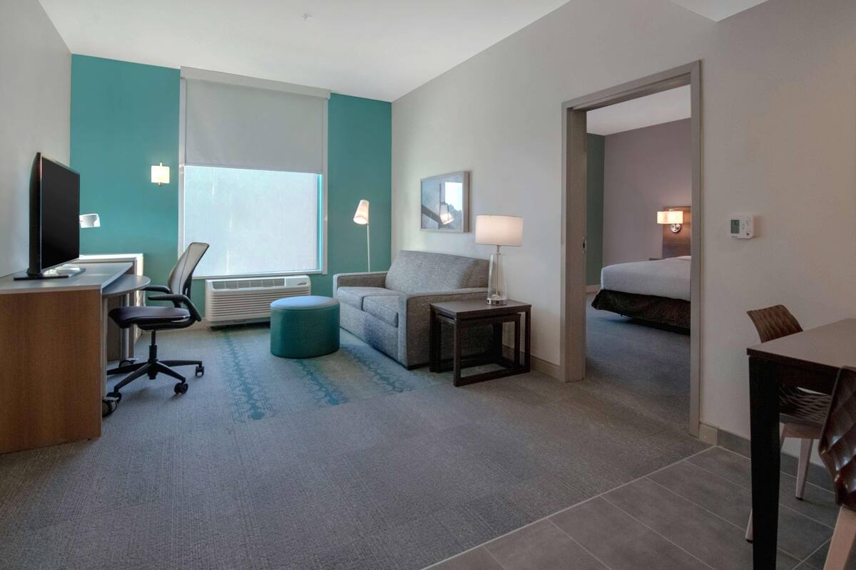Habitació d'hotel amb escriptori, cadira, sofà, otomana, taulella de nit, làmpada i llit.