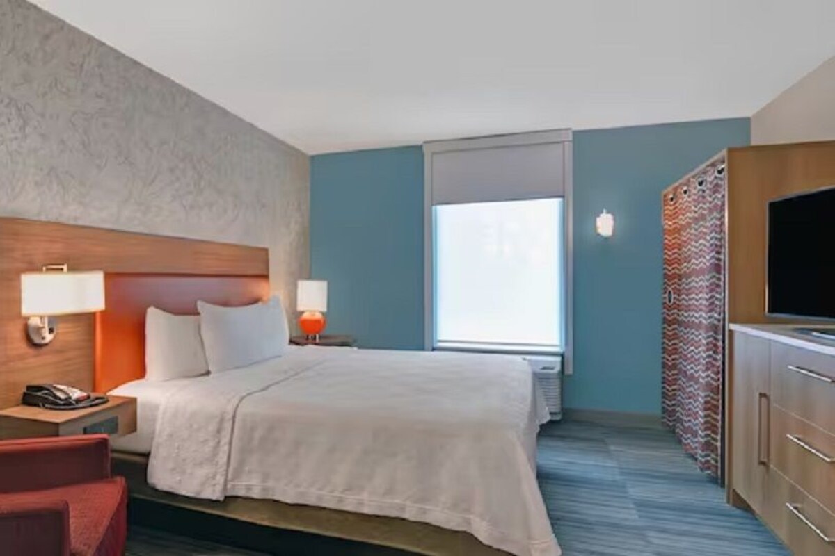 Une chambre d’hôtel avec un lit, un fauteuil rouge, une télévision et une fenêtre avec des rideaux.