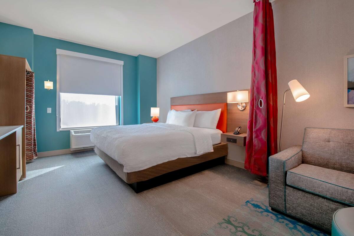 Habitació d'hotel amb un llit, taules de nit, una cadira i una finestra amb cortines.