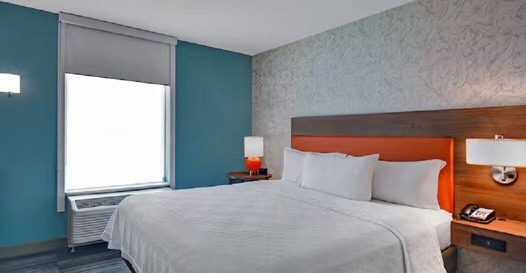 A hotel room with a large bed, a wooden headboard, a nightstand with a lamp, and a window with blinds.