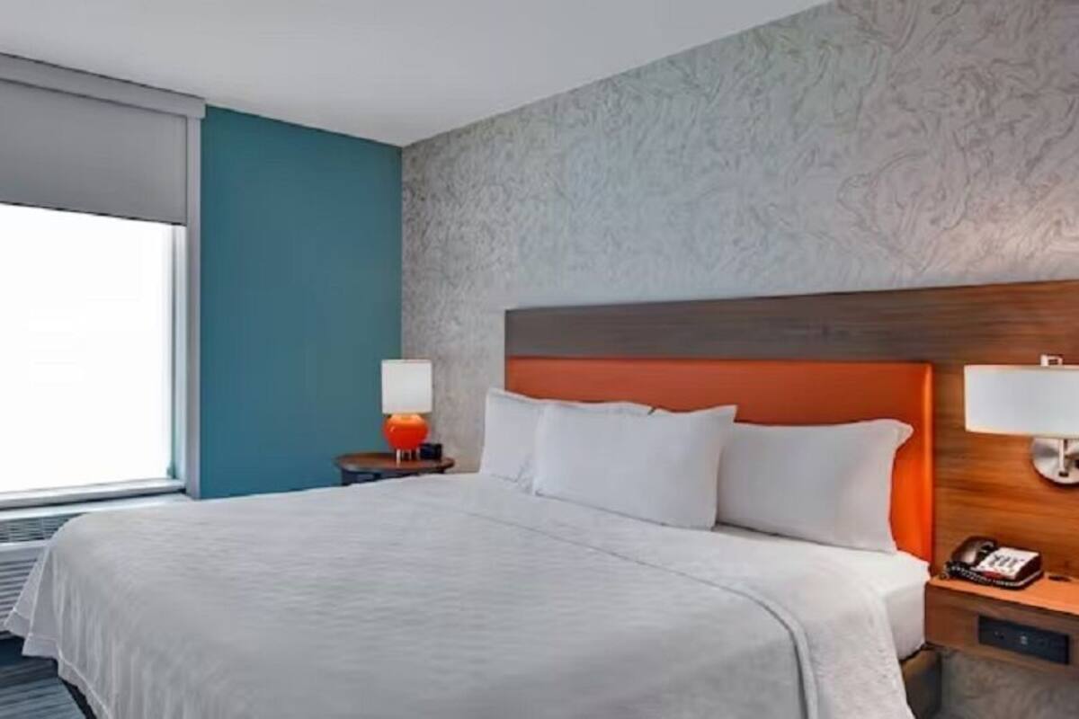 Habitació d'hotel amb un llit gran, un capçal de fusta, una nitzeta amb una làmpada i una finestra amb persianes.