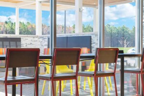 Un área de estar moderna al aire libre con paredes de vidrio, sillas rojas y vistas a árboles y un estacionamiento.