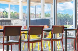 Un área de estar moderna al aire libre con paredes de vidrio, sillas rojas y vistas a árboles y un estacionamiento.