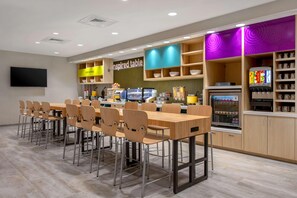 Un restaurant moderne avec une longue table et des chaises en bois, un espace cuisine équipé d’une machine à café, et une enseigne sur laquelle on peut lire : « Table d’inspiration ».