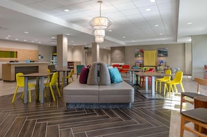 Un moderno vestíbulo de hotel con un área de descanso central, un mostrador de recepción y un comedor con sillas amarillas.
