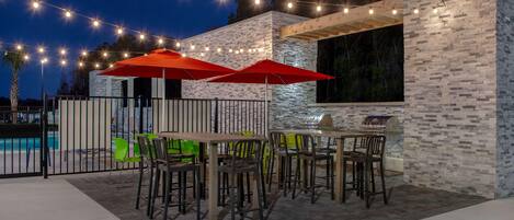 Un patio al aire libre con mesas y sillas, luces colgantes y una piscina al fondo.
