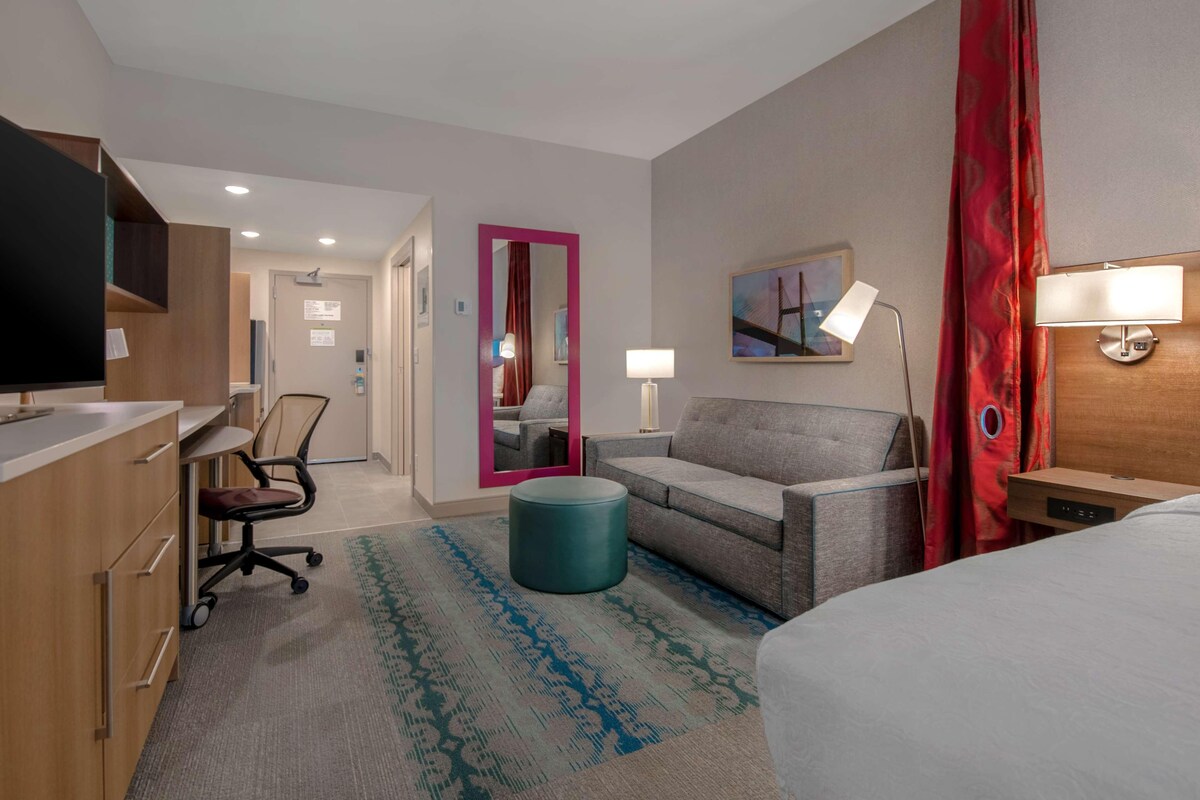 Habitación de hotel con cama, escritorio, silla, sofá, espejo y lámpara.