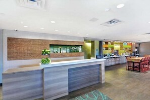 A modern hotel reception area with a wooden accent wall, a reception desk, and a dining area with red chairs. - Home2 Suites By Hilton Savannah I95 North (Port Wentworth)