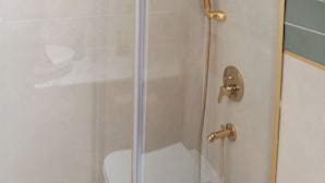 A glass-enclosed shower with a gold showerhead and handheld showerhead, a glass door, and a white tiled wall. - Taksim Bosphorus Hotel (Istanbul)