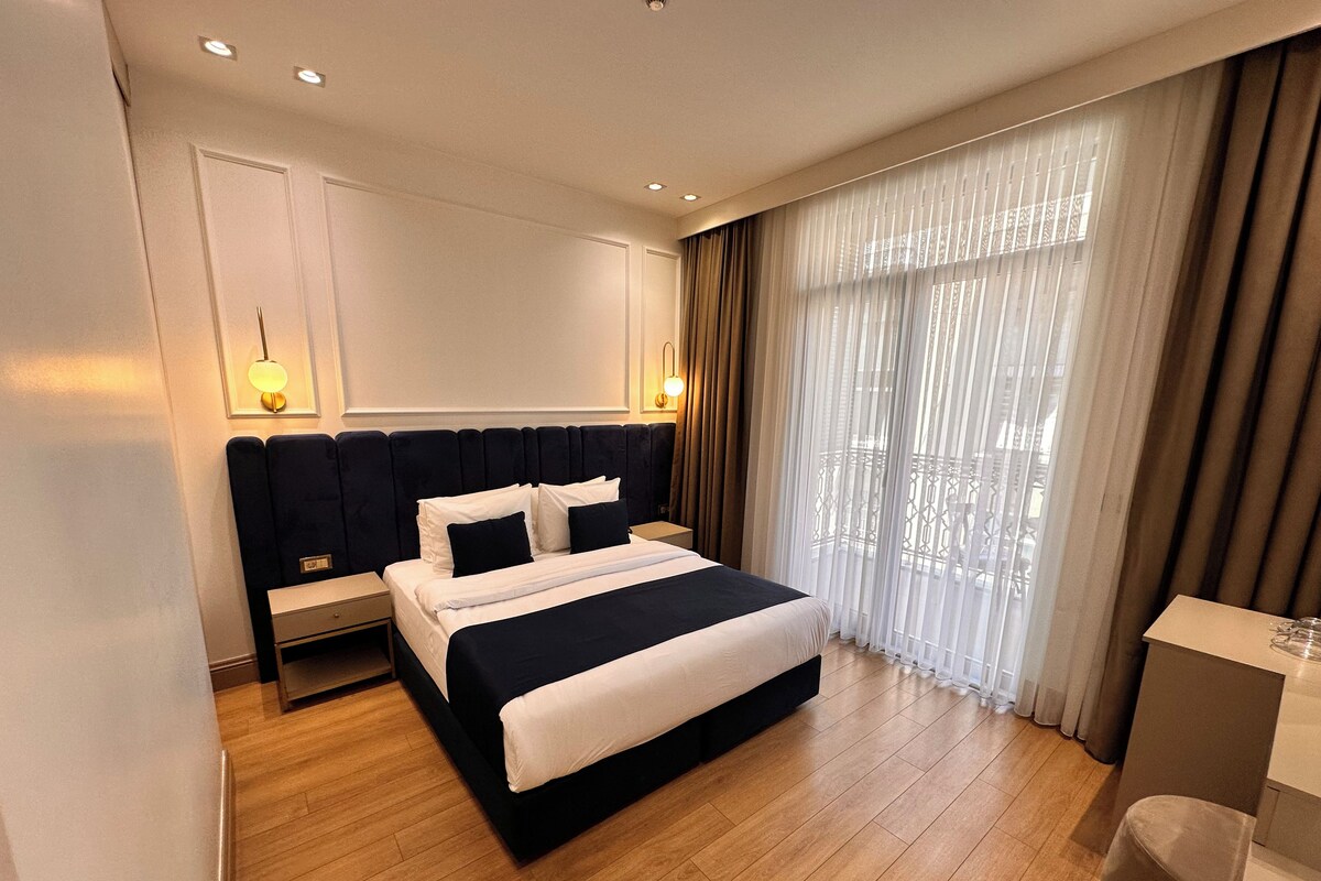 A modern hotel room with a large bed, a bedside table, a lamp, and a view of the outdoors through sheer curtains.