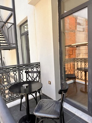 Un balcó amb taula i caires, una barana metàl·lica i vista d'un edifici de maó a través d'una porta de vidre.