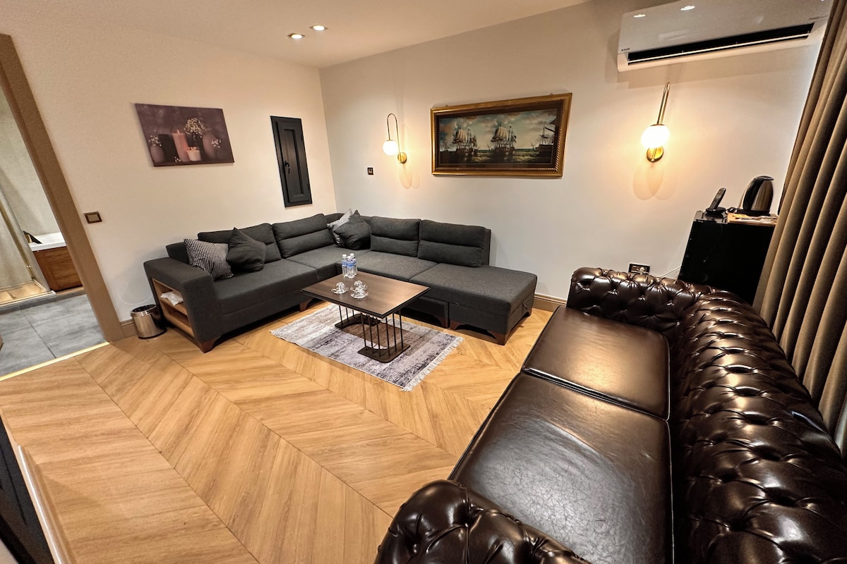 A modern living room with a sectional sofa, a coffee table, and a leather ottoman. There are wall-mounted lights and a painting above the sofa.