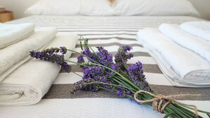 Ein ordentlich bezogenes Bett mit weißen Leinen und einer Streifen-Decke, ein Strauß Lavendelblumen, gebunden mit einer Schnur, und gefaltete Handtücher auf dem Bett.