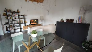 Ein modernes Wohnzimmer mit einem Glastisch, weißen Stühlen, einem Bücherregal, einem Fernseher und einem schwarzen Schrank.