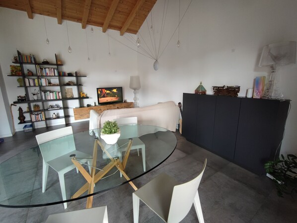 A modern living room with a glass dining table, white chairs, a bookshelf, a television, and a black cabinet.