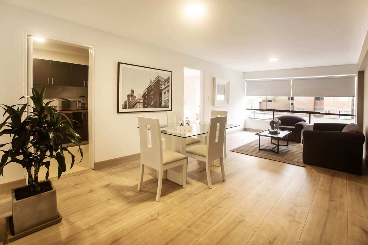 Ein modernes Wohnzimmer mit einem Glastisch, weißen Stühlen, einem dunklen Sofa und einem großen Fenster mit Jalousien.