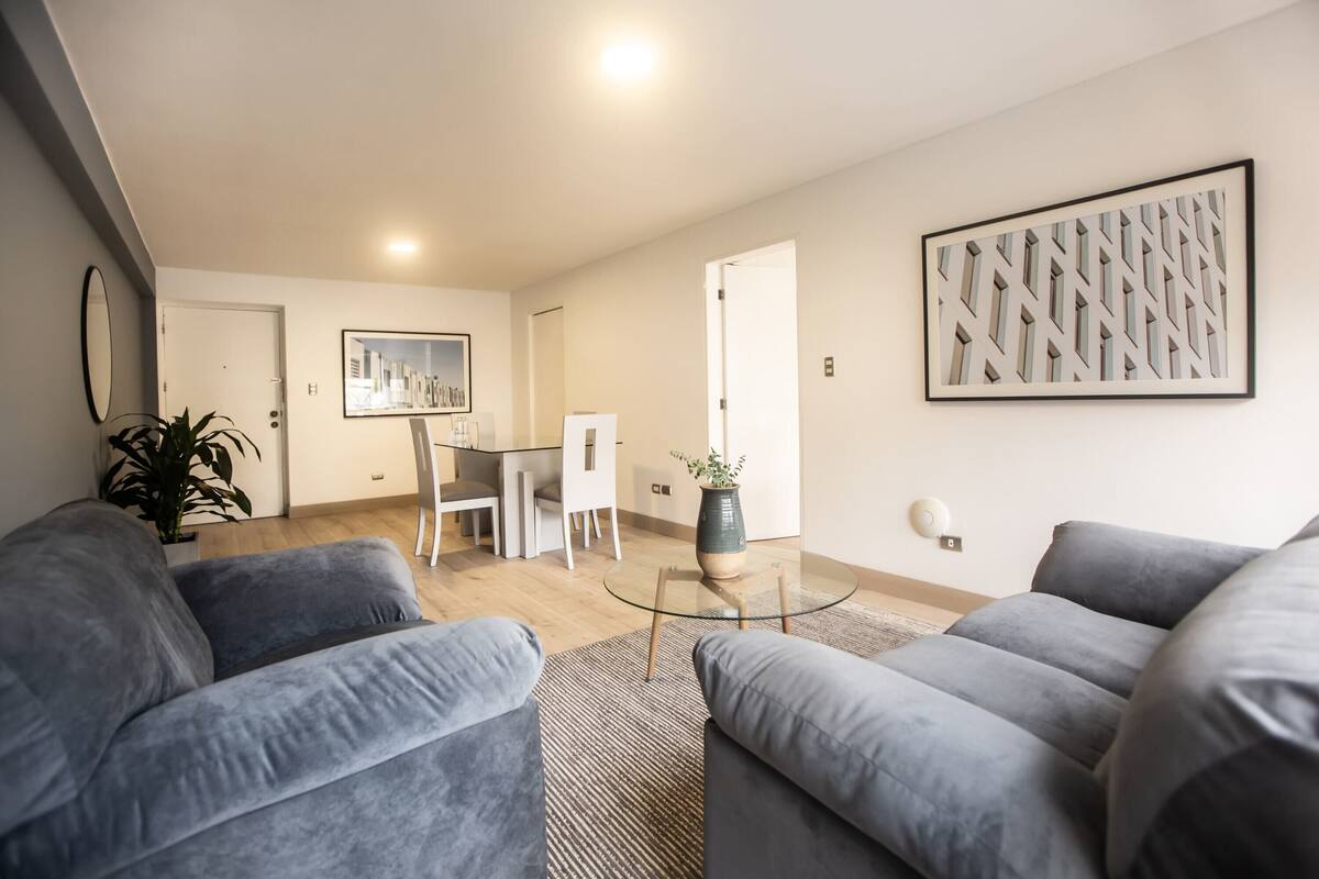 Ein modernes Wohnzimmer mit grauen Sofas, einem Glastisch und einem gerahmten abstrakten Kunstwerk an der Wand.