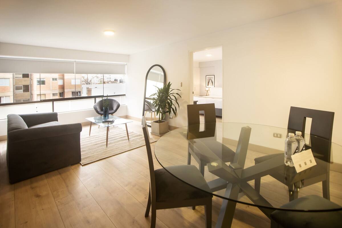 Ein modernes Wohnzimmer mit einem Glastisch, dunklen Sofas und einem großen Fenster mit Blick auf Gebäude.