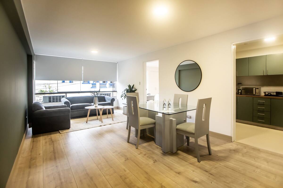 Ein modernes Wohnzimmer mit einem Glastisch, grauen Stühlen, einem grauen Sofa und einem kleinen Beistelltisch. Rechts ist eine Küche mit grünen Schränken und einer Mikrowelle erkennbar.