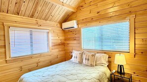 A wooden cabin bedroom with a bed, two windows with blinds, an air conditioning unit, and a bedside table with a lamp. - Cozy Cabin with Private Resort Amenities: Walking Distance to Lake James (Marion)