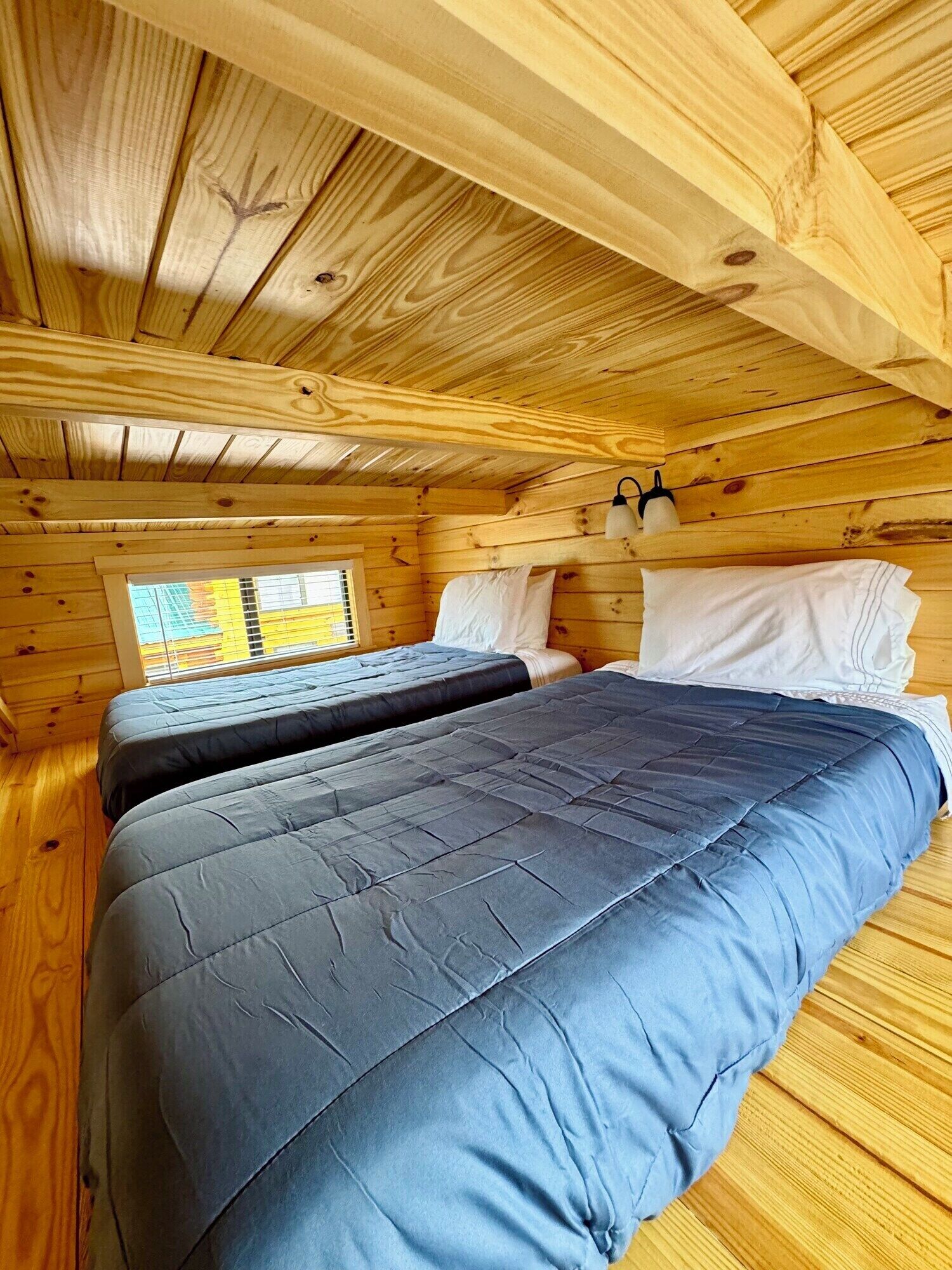 A wooden cabin room with two beds, a window, and a wall-mounted lamp.