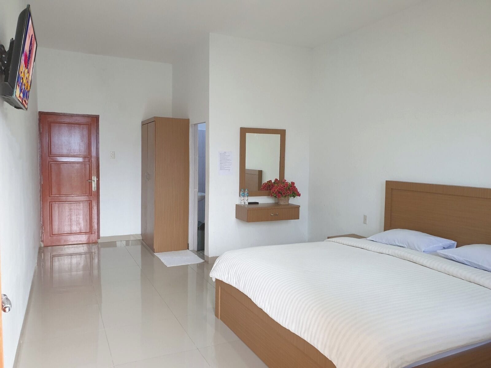 Um quarto com cama, guarda-roupa de madeira, espelho e uma mesinha com um vaso de flores.