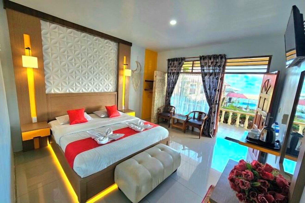 A modern hotel room with a large bed, a bedside table, a nightstand, a seating area with a chair and table, and a view of a pool.