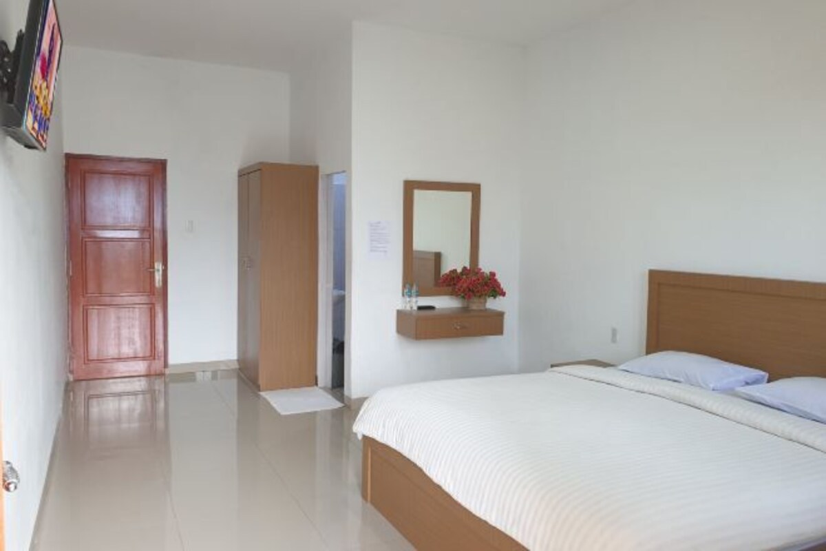 A hotel room with a large bed, a wooden wardrobe, a mirror, and a small table with a vase of flowers.