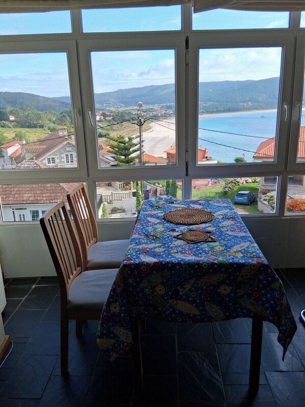 Comedor con una mesa cubierta con mantel estampado, una silla de madera y vistas a un paisaje costero a través de amplios ventanales.