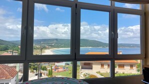 A room with a large window overlooking a beach, hills, and a small village. - The apartment is located in front of Langosteira beach, with stunning views. (Fisterra)