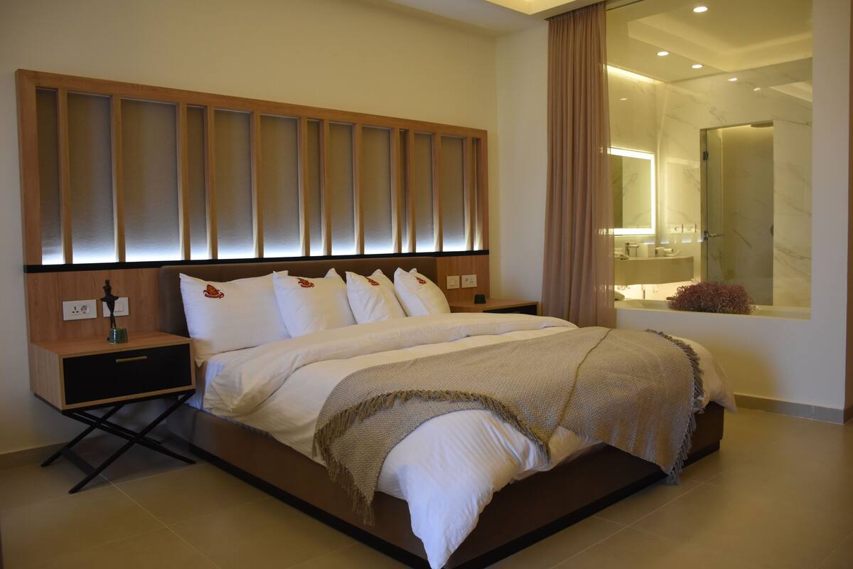 A modern hotel room with a large bed, a nightstand, a mirror, and a bathroom visible through a glass door.