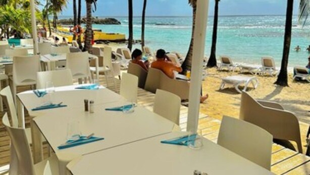 An outdoor dining area with white tables and chairs, a beach view, and palm trees. - Studio Relax Manganao, Proche de la plage et accès a la piscine (Saint-François)