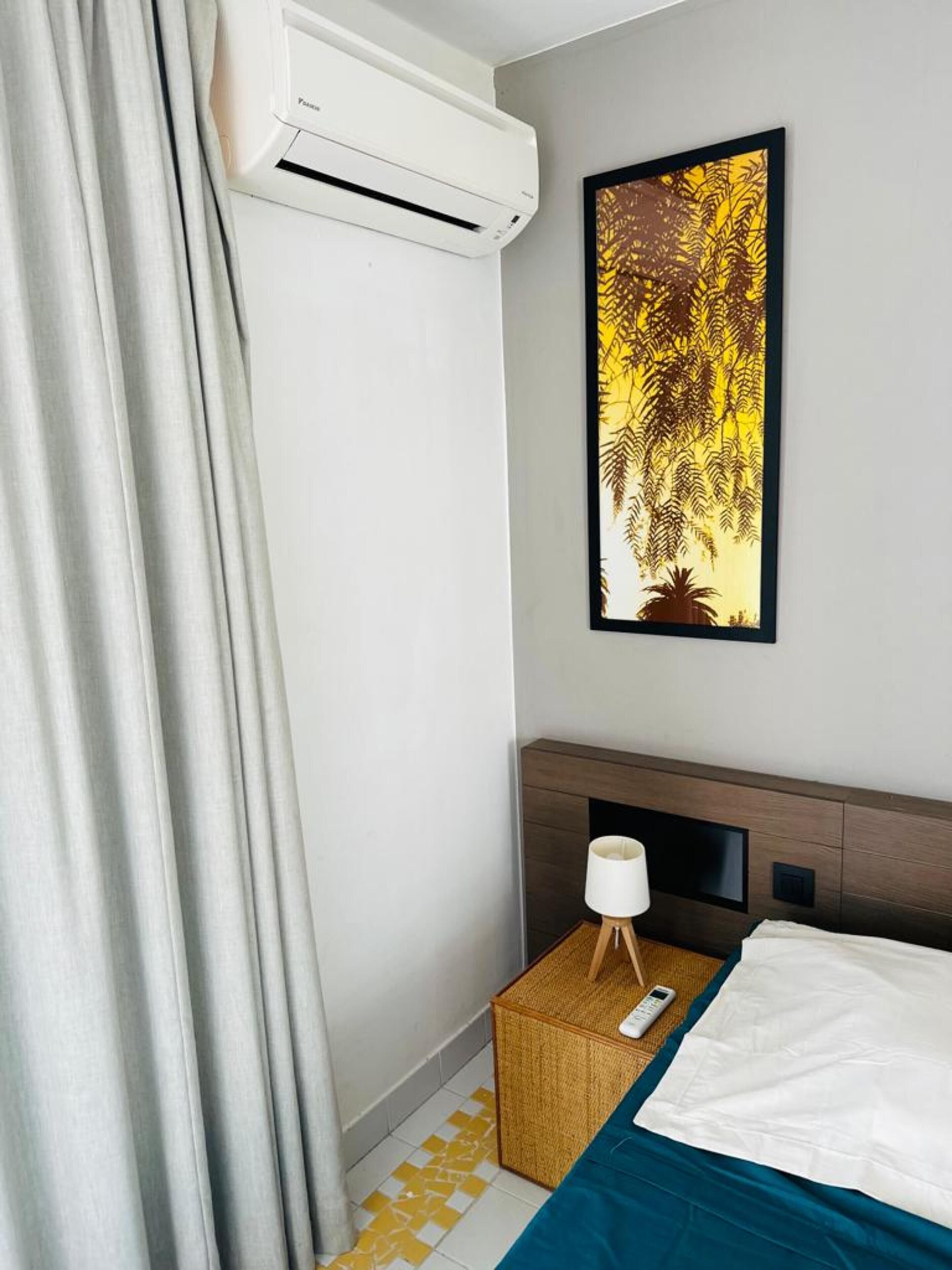 A hotel room with a bed, a bedside table with a lamp, a wall-mounted air conditioner, and a framed artwork.