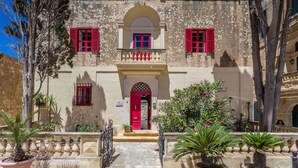 Un bâtiment historique avec une porte et des fenêtres rouges, entouré de plantes en pot et d’arbres.