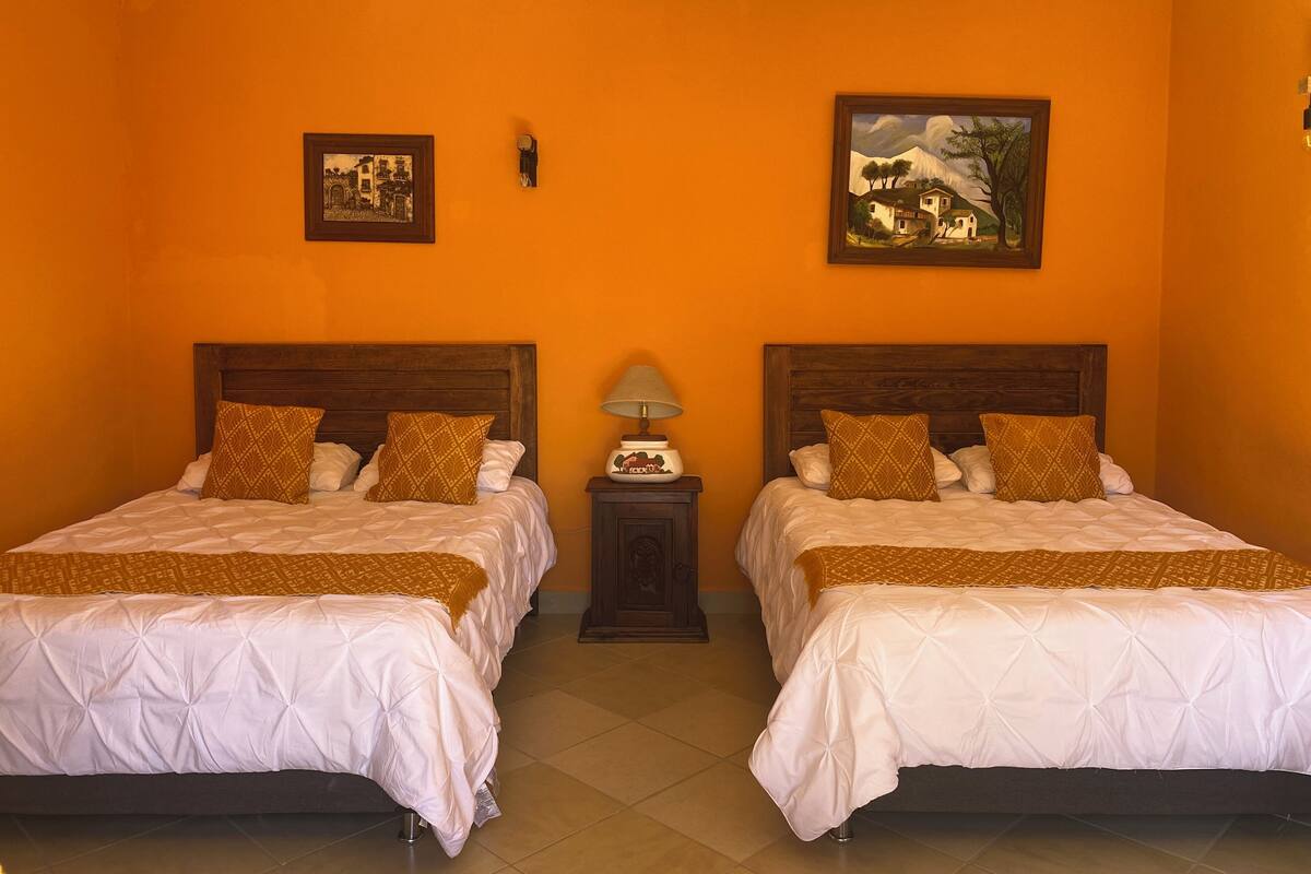 A hotel room with two beds, each with a wooden headboard, white bedding, and patterned pillows. There are two framed pictures on the walls, a nightstand with a lamp, and a ceiling fan.